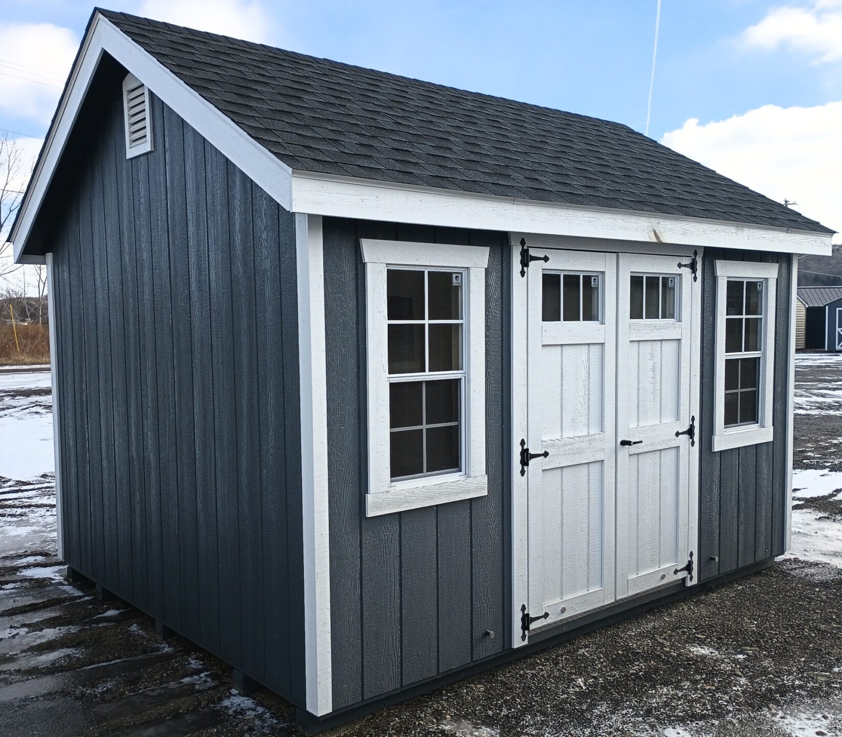 Dark gray building with white trim, two windows, shingled roof and double doors