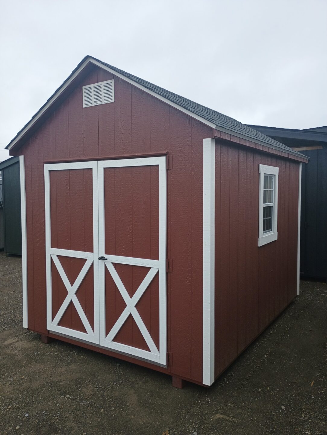 Small red shed with white trim, double doors, gable vents, windows on either side and a shingled roof